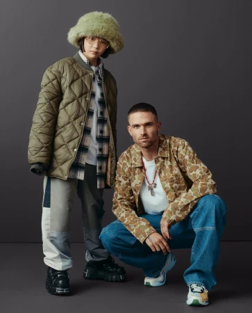 Two stylish individuals posing in a studio against a grey background. One is standing wearing a green coat and the other is crouching.