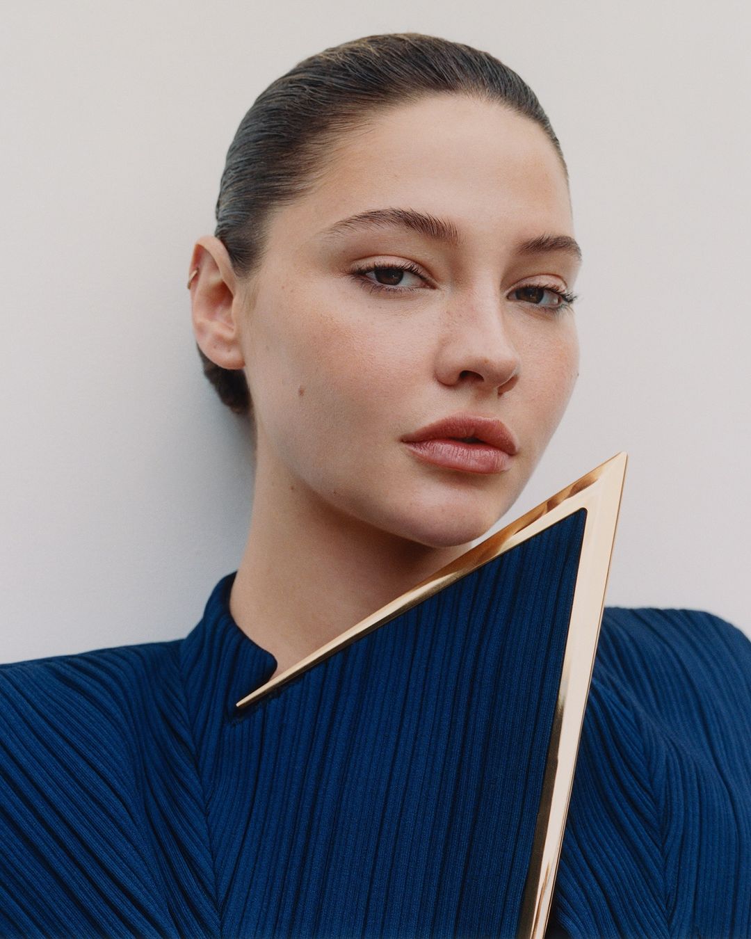 Close-up studio portrait of a young woman with dark hair and neutral makeup, wearing a blue dress and holding a golden, triangular object.