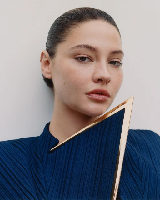 Close-up studio portrait of a young woman with dark hair and neutral makeup, wearing a blue dress and holding a golden, triangular object.