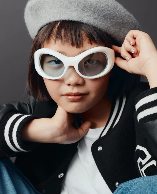 Young child in a beret and oversized sunglasses posing indoors. The subject is a medium shot against a grey background.