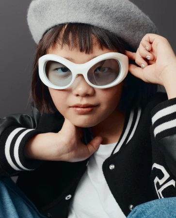 Young child wearing oversized white sunglasses, grey beret, and black varsity jacket against a grey background