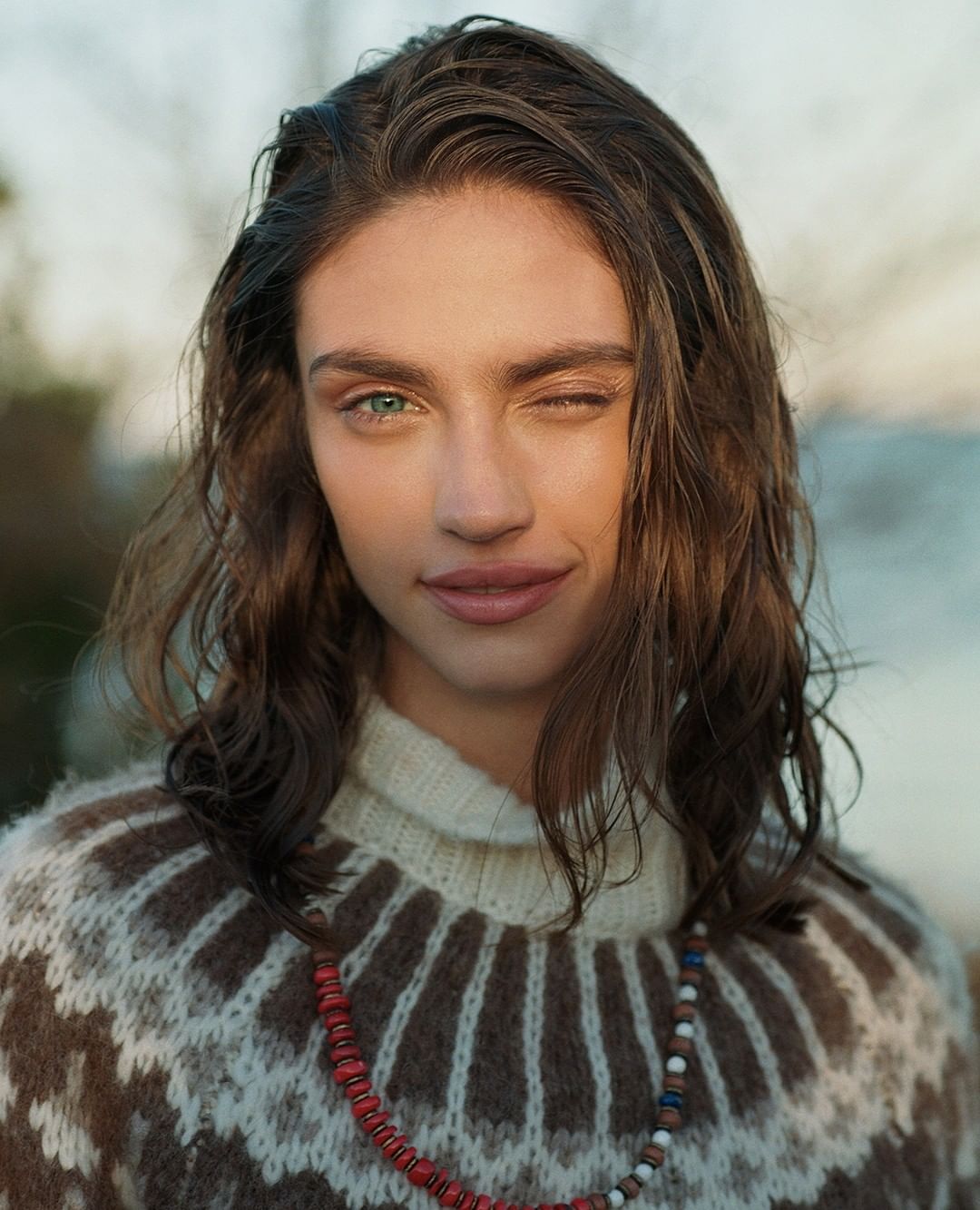 Close-up portrait of a young woman with heterochromia wearing a patterned sweater and beaded necklace, set against a natural outdoor backdrop.