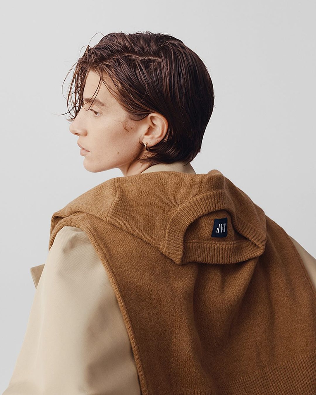 Profile shot of a young adult with short brown hair wearing a brown sweater and a light-brown overcoat on a neutral background.