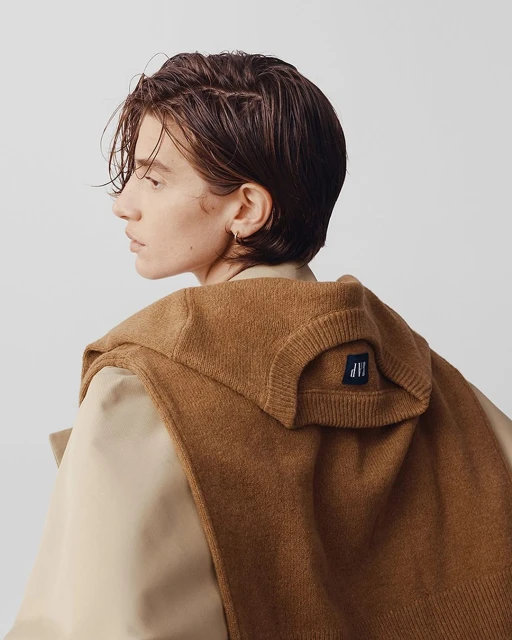 Profile shot of a young adult with short brown hair wearing a brown sweater and a light-brown overcoat on a neutral background.