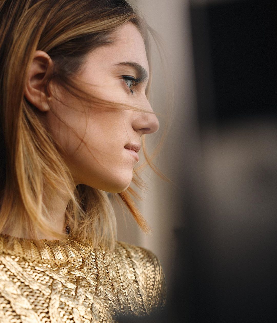 Profile shot of a woman with shoulder-length blonde hair wearing a gold, textured sweater against a blurred gray background.