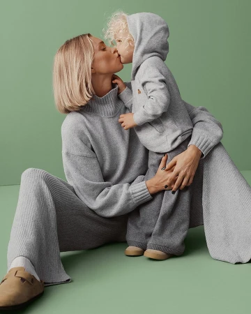 Blonde woman and curly-haired toddler share a kiss, both wearing gray knitwear, seated on a green studio backdrop.