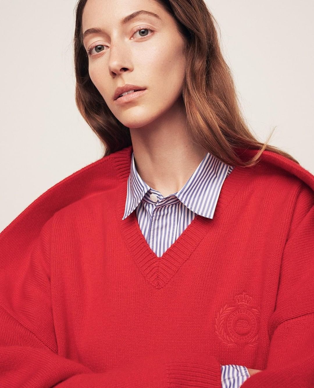 Portrait of a fair-skinned adult woman wearing a red V-neck sweater over a blue and white striped shirt, posing indoors.