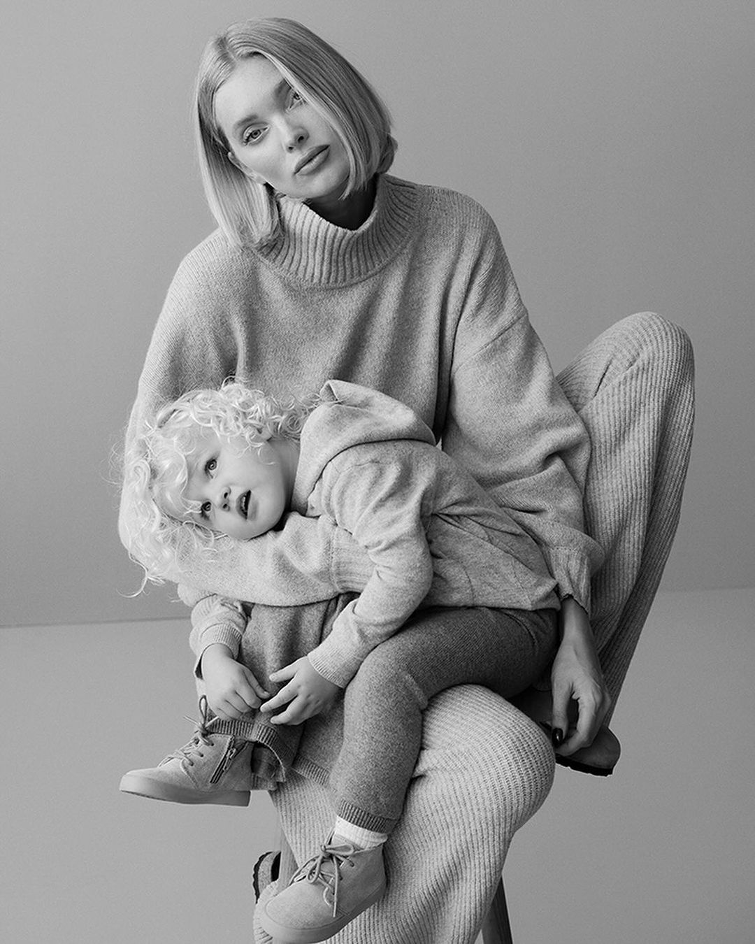 A black and white portrait of a mother in a knit outfit holding her child who is wearing matching shoes in a studio setting.
