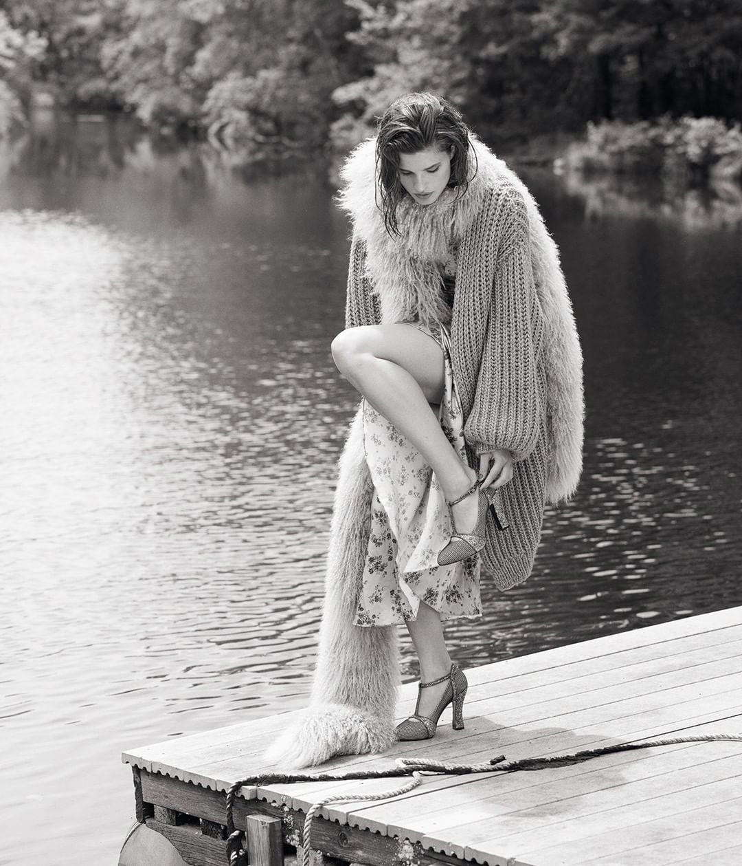 Model on a wooden dock wearing a furry knit cardigan adjusting high heel sandals by a body of water