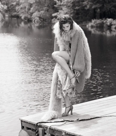 Model on a wooden dock wearing a furry knit cardigan adjusting high heel sandals by a body of water
