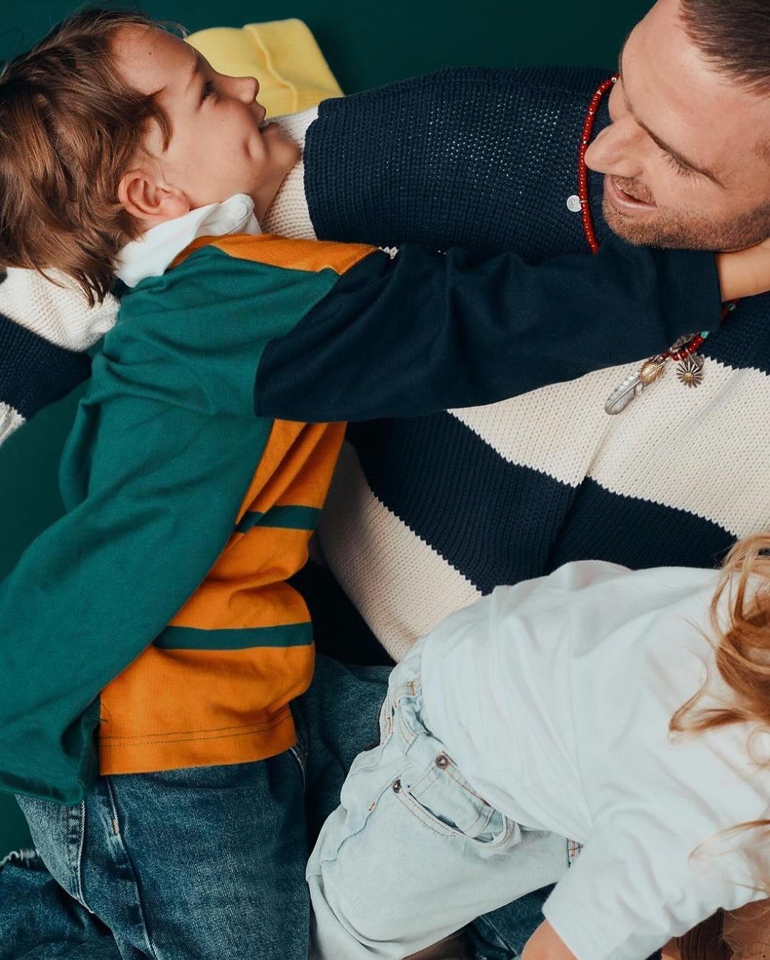 A joyful moment captured indoors shows a dad and two kids in a playful embrace. They are wearing casual clothing, and the atmosphere is warm and loving.