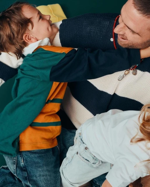 A joyful moment captured indoors shows a dad and two kids in a playful embrace. They are wearing casual clothing, and the atmosphere is warm and loving.