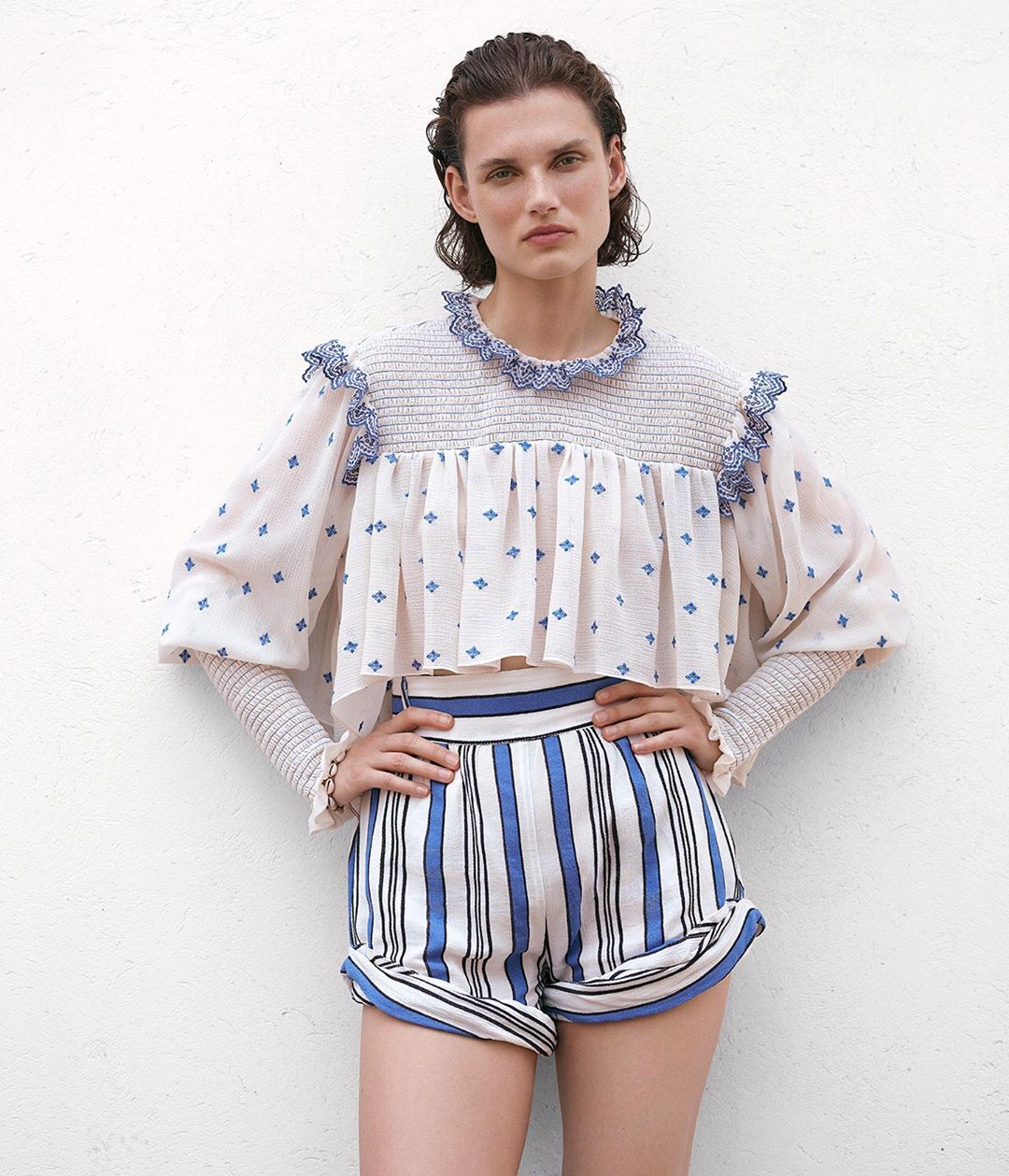 Fashion model in a photo studio wearing a white smocked ruffle top with blue diamond pattern, and high-waisted striped shorts.