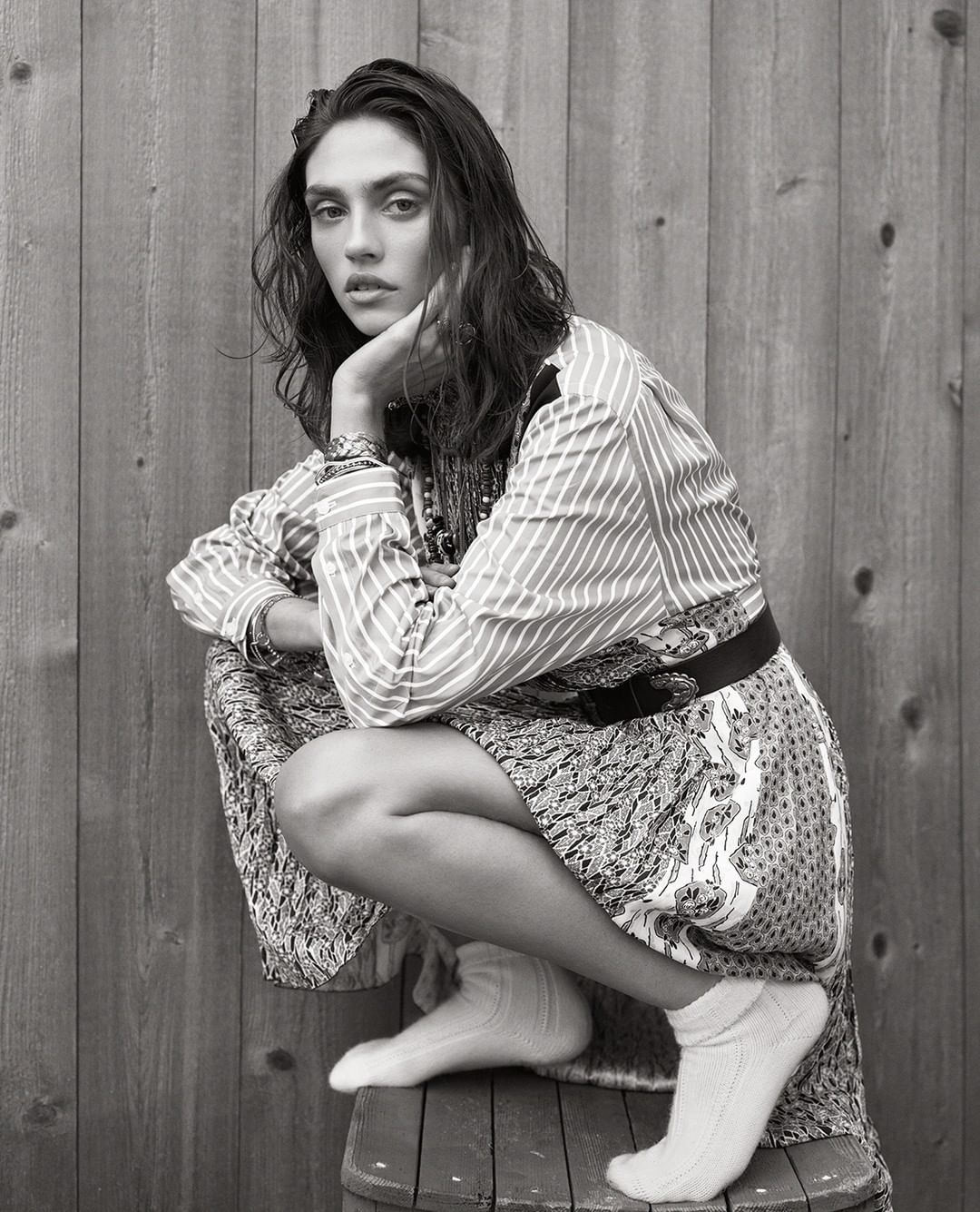 Portrait of a woman with long dark hair, wearing a patterned dress and white socks, sitting on a barrel in front of a wooden wall.