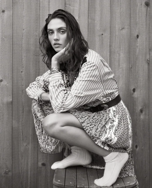 Portrait of a woman with long dark hair, wearing a patterned dress and white socks, sitting on a barrel in front of a wooden wall.