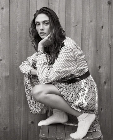 Portrait of a woman with long dark hair, wearing a patterned dress and white socks, sitting on a barrel in front of a wooden wall.