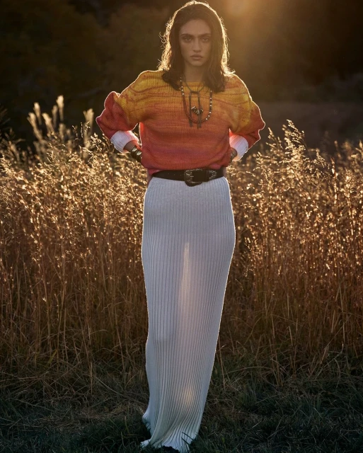 A woman in a sweater and skirt poses outdoors in a field of tall grass at sunset. Fashion editorial shot with natural light.