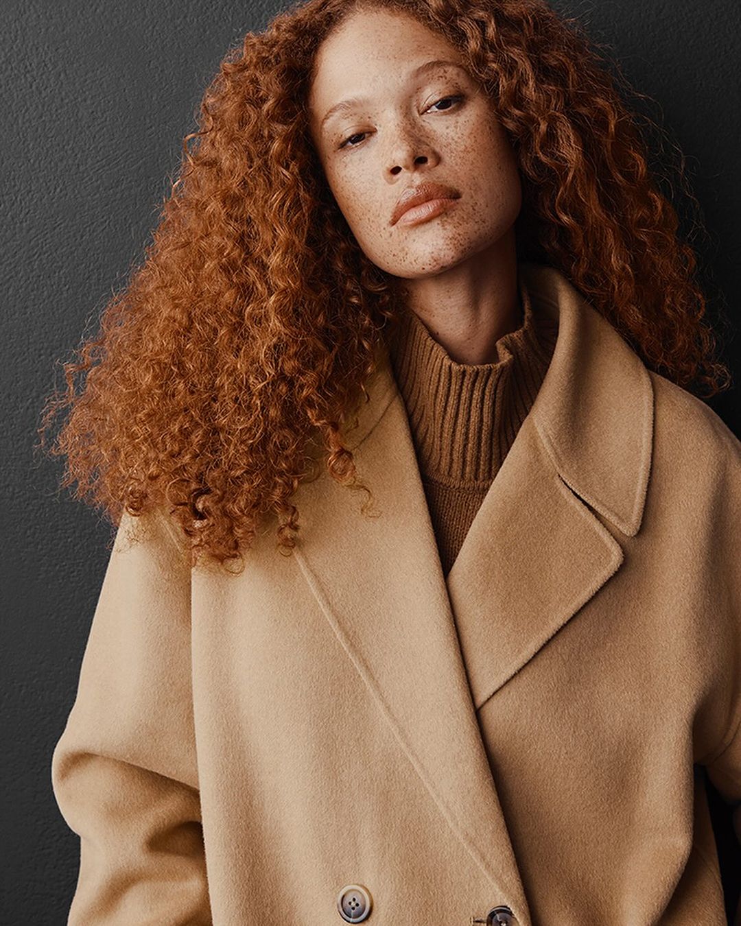 Close-up of a red-haired woman in a tan wool coat with a turtleneck, captured in a studio with a dark background, fashion portrait.