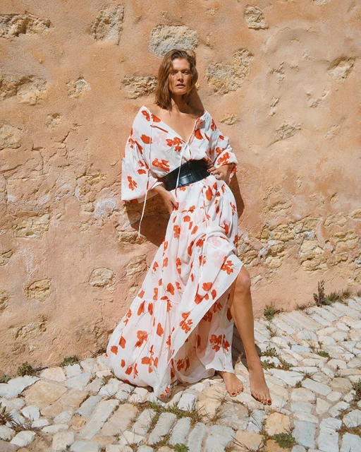 A woman in a white, floral-print dress and wide black belt poses in front of a textured wall and cobblestone in warm, natural light.