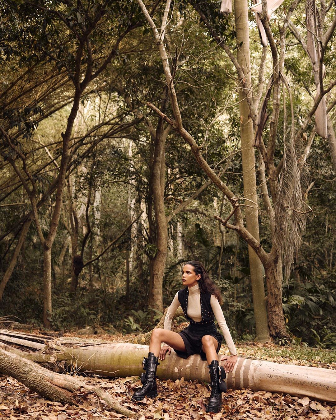 Woman in dark miniskirt and boots sits on fallen tree trunk in forest setting with tall trees and leafy ground cover.