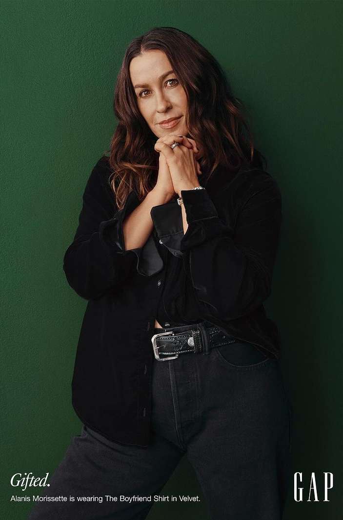 Alanis Morissette poses in a velvet shirt and jeans against a green background for GAP. She has her hands clasped together.