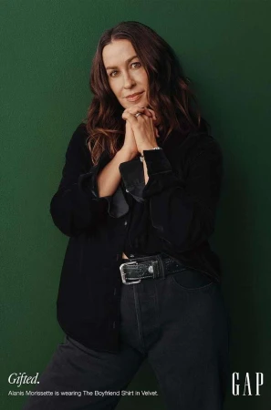 Alanis Morissette poses in a velvet shirt and jeans against a green background for GAP. She has her hands clasped together.