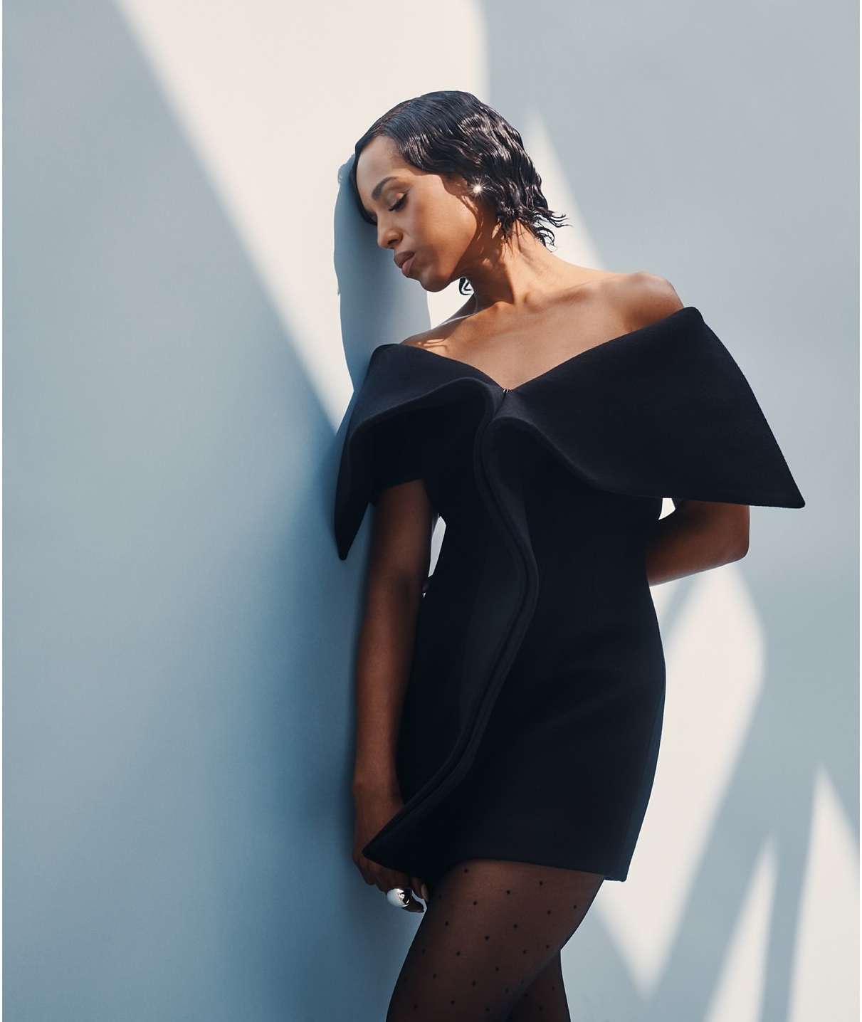 Elegant woman in a short black off-the-shoulder dress with a large bow poses against a light blue wall with bright, soft lighting.