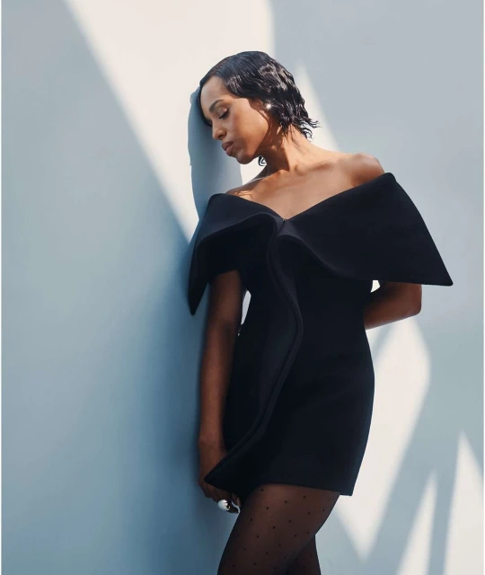 Elegant woman in a short black off-the-shoulder dress with a large bow poses against a light blue wall with bright, soft lighting.