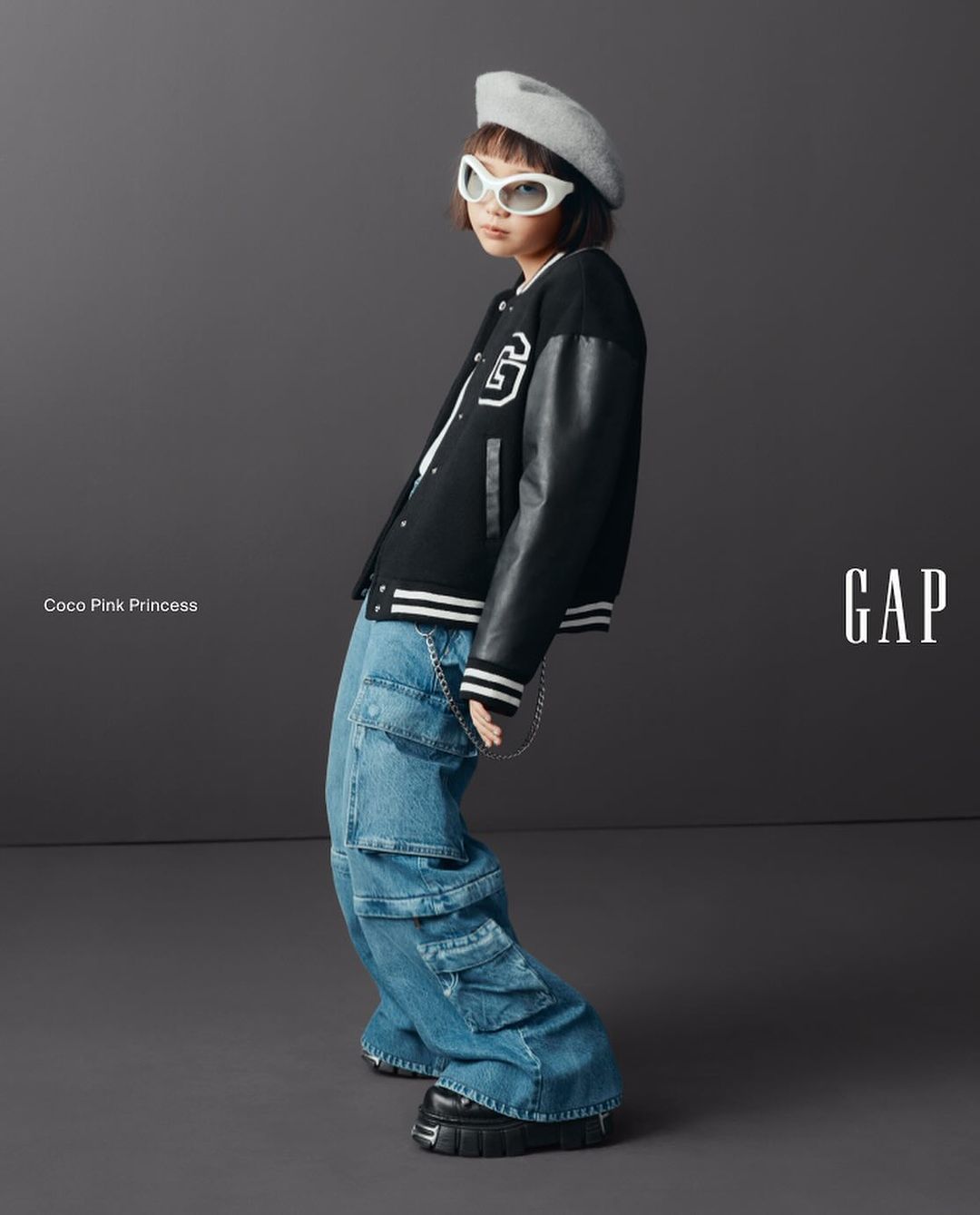Child fashion model in a black varsity jacket, baggy jeans, and white sunglasses, standing in a studio against a gray background.