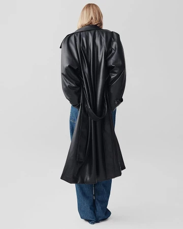 Model seen from behind wearing a long black leather trench coat and blue jeans against a white seamless background
