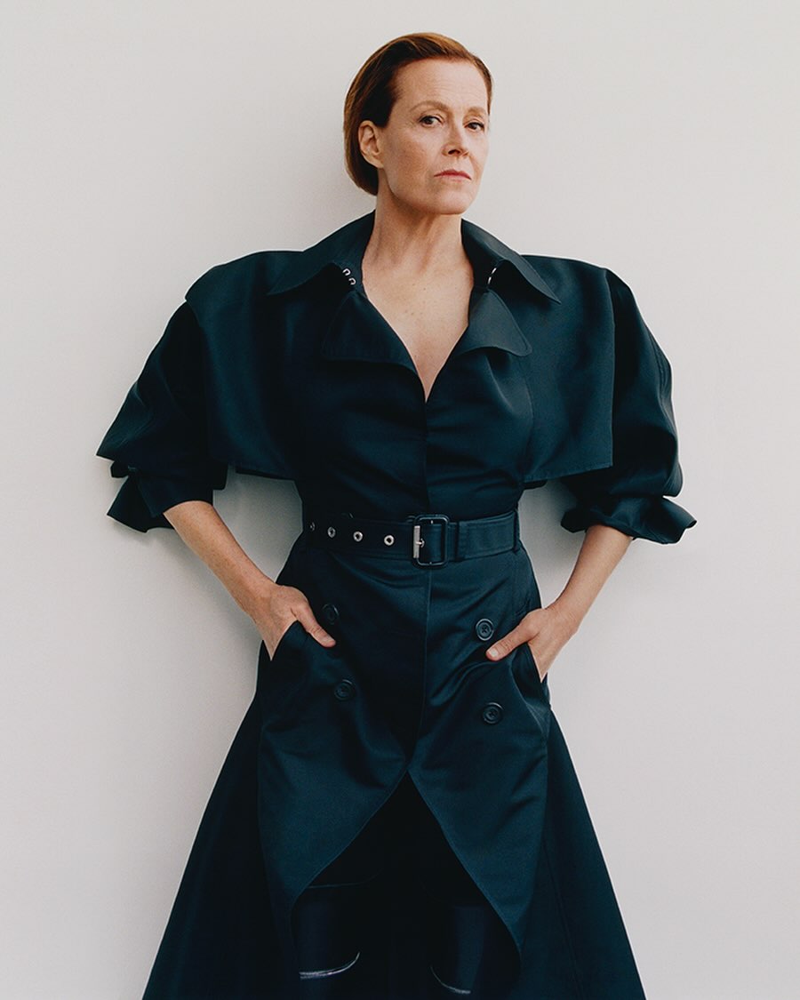 Elegant woman in a belted black dress stands indoors against a white wall, exuding confidence and sophistication in a studio portrait.