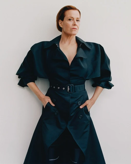 Elegant woman in a belted black dress stands indoors against a white wall, exuding confidence and sophistication in a studio portrait.