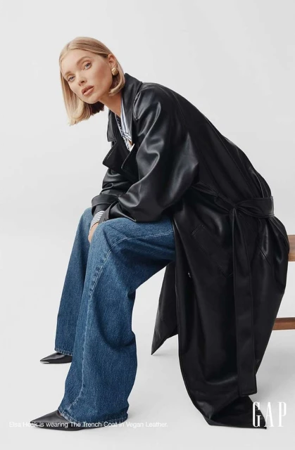 Fashion shot of a blonde woman in a long black coat and blue jeans sitting on a wood stool in a white studio setting.