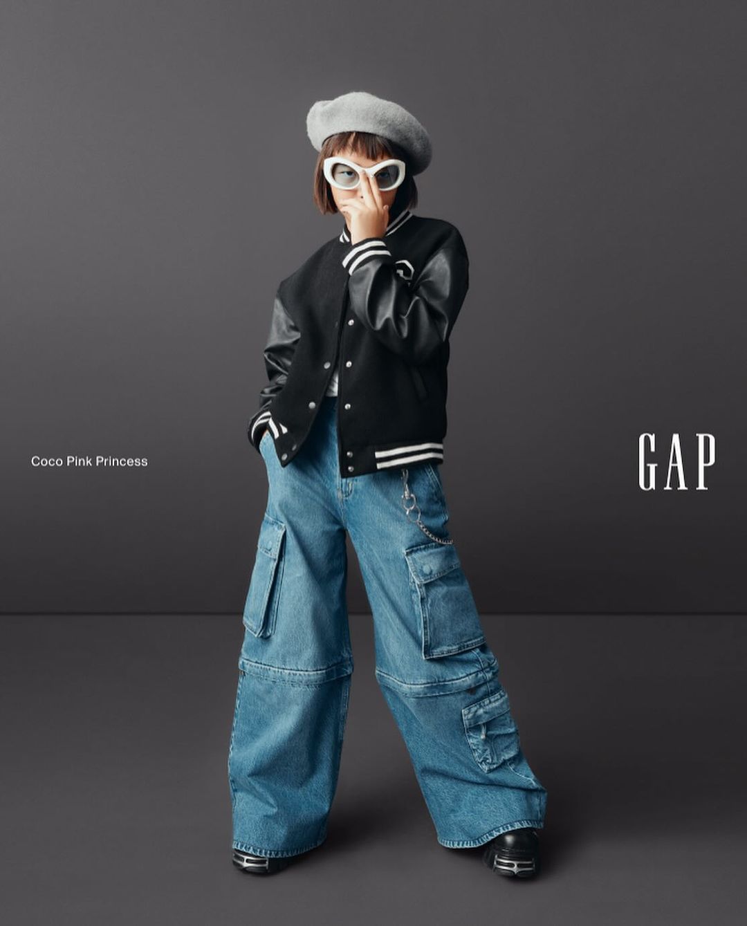 Young girl in stylish streetwear fashion poses in a studio wearing a beret, letterman jacket, and baggy jeans for a Gap ad.