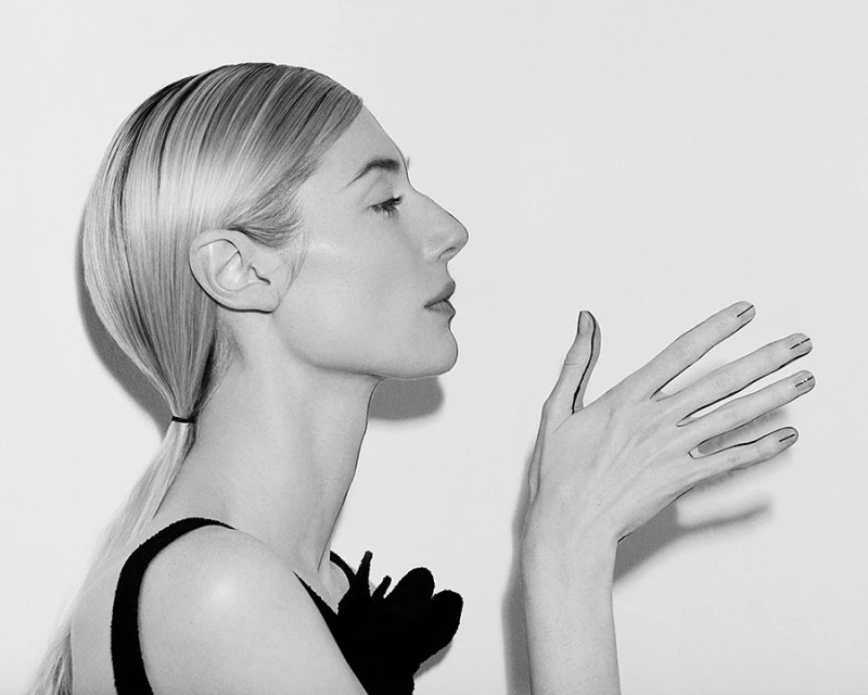 Profile view of an adult woman with blonde hair in a ponytail, wearing a black top and a flower, with her hand raised indoors.