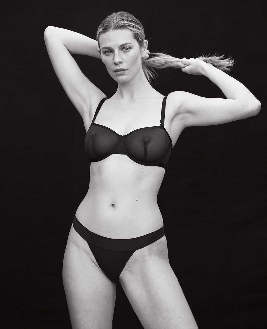 Monochrome portrait of a woman in black underwear adjusting her hair against a dark background