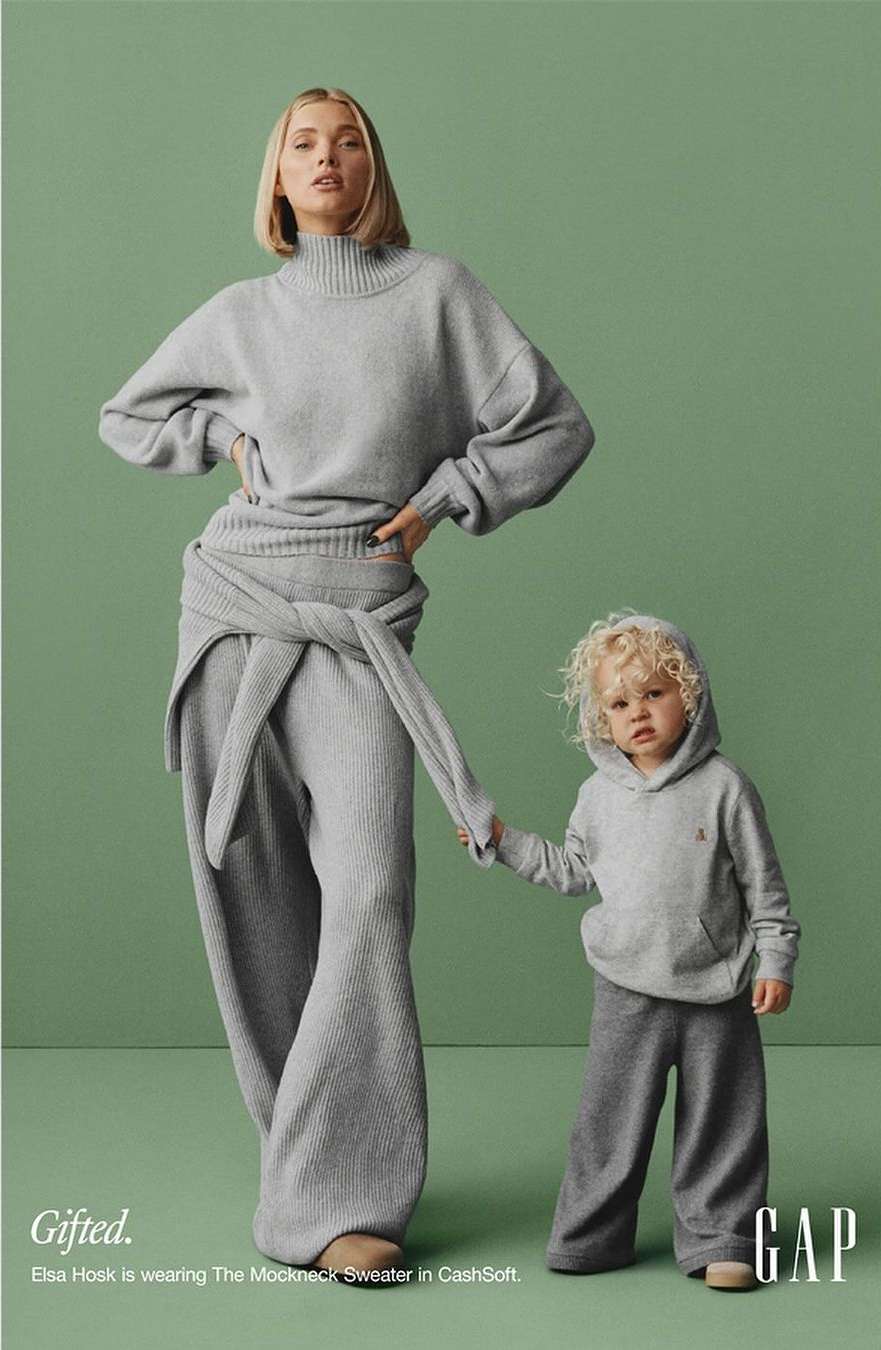 A blonde adult female and blonde toddler holding hands wearing grey clothing on a green background in a GAP advertisement.