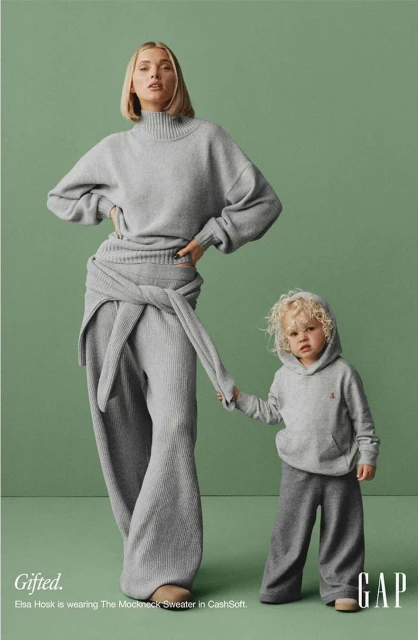 A blonde adult female and blonde toddler holding hands wearing grey clothing on a green background in a GAP advertisement.