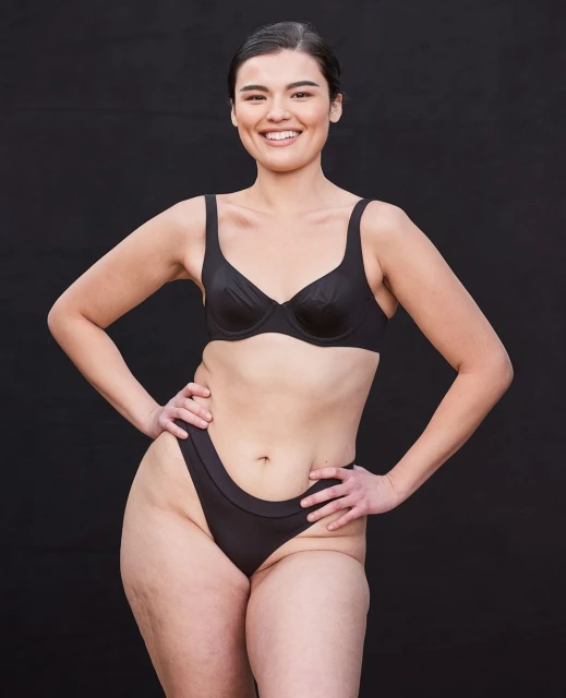 Smiling woman in black bra and underwear poses against a dark background, showcasing a body-positive image in a studio setting.