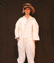 Model in white jumpsuit and straw bucket hat standing against a textured dark backdrop in New York by Bjorn Iooss.