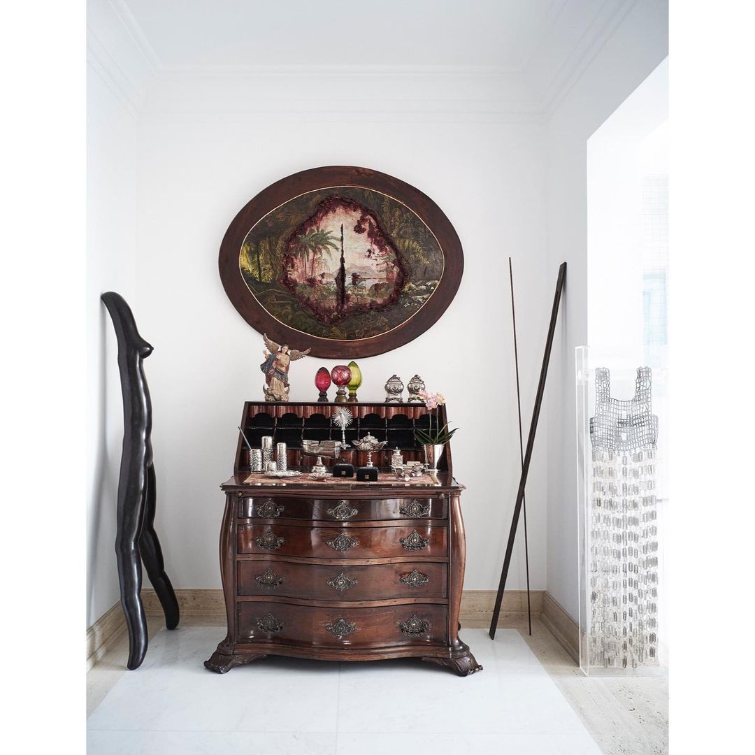 Elegant interior featuring an antique wooden secretary desk, oval painting, and sculptural elements against a minimalist white wall backdrop.