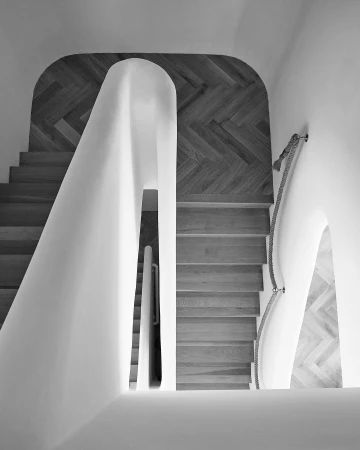 High-angle shot of a modern interior with wooden stairs, white walls, and a herringbone wood pattern on the upper landing, in black and white.
