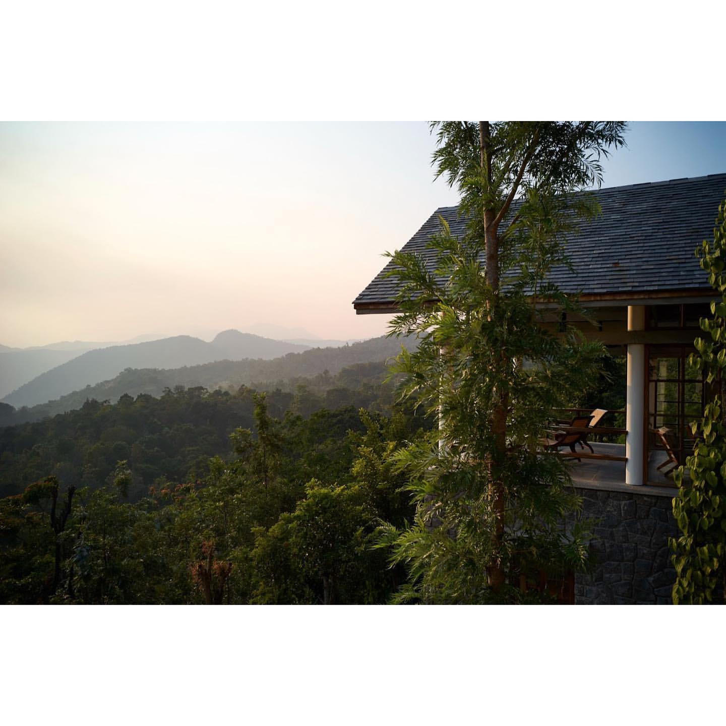 Scenic view of a hillside house exterior featuring a gray roof, stone pillar supports, outdoor seating, and a lush green forest landscape.
