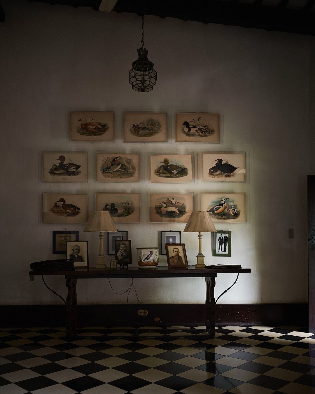 Interior view of a room featuring framed prints of birds, a table with lamps, and a black and white checkered floor, evoking a vintage ambiance.