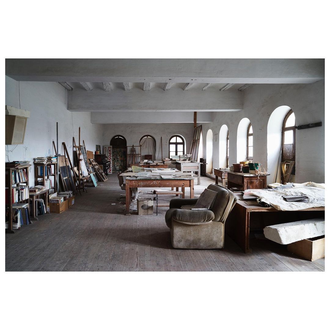 Spacious artist studio filled with work tables, easels, books, and a vintage armchair, illuminated by natural light through arched windows.