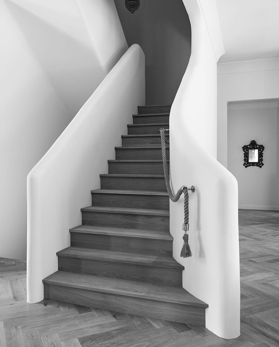 A black and white interior shot of a staircase featuring wood steps, rope railing, and white walls with herringbone-patterned flooring.