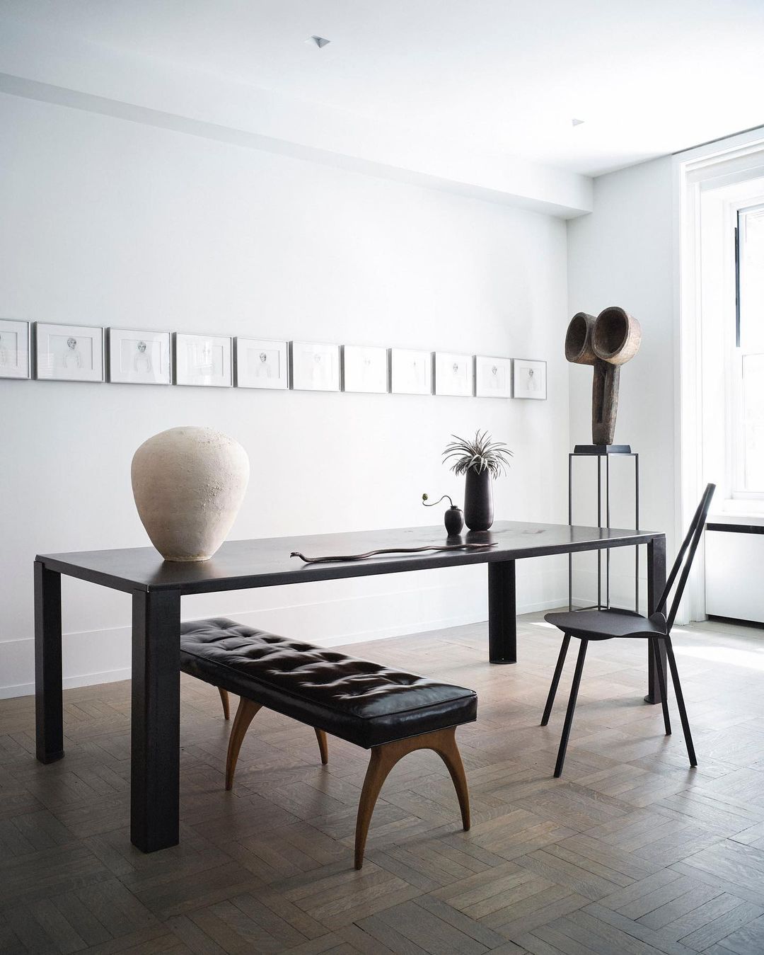 Interior photo of a black dining table with a bench and chair, a vase, sculpture, and framed art in a minimalist white dining room.