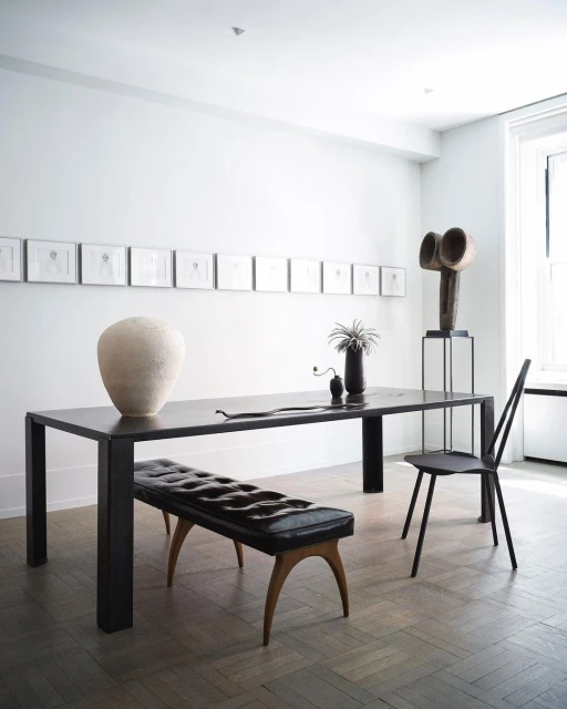 Interior photo of a black dining table with a bench and chair, a vase, sculpture, and framed art in a minimalist white dining room.