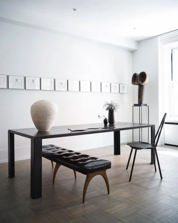 Interior photo of a black dining table with a bench and chair, a vase, sculpture, and framed art in a minimalist white dining room.