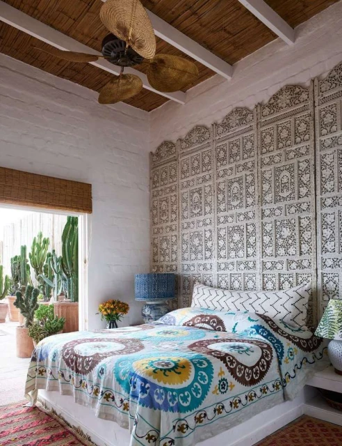 Bedroom interior featuring a bed with a patterned duvet cover, an ornate headboard, and a view of cacti in a bright, airy space.